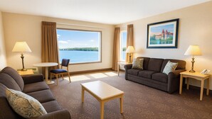 A hotel room with a sofa, armchair, and a small table. There is a large window with a view of a body of water and a building in the distance. - Bridge Vista Beach - Hotel & Convention Center (Mackinaw City)
