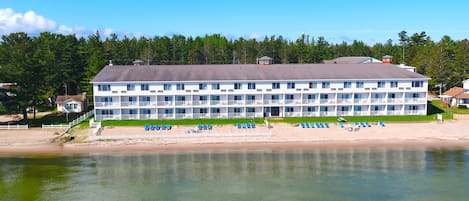 Ein mehrstöckiges Hotel mit flachem Dach, in Strandnähe, mit blauen Stühlen und Sonnenschirmen.