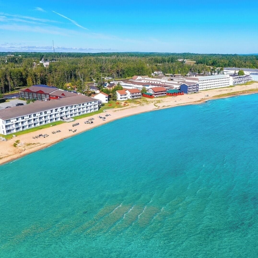Vue aérienne d’un hôtel situé sur la côte, composé de plusieurs bâtiments, avec une plage de sable et une eau turquoise cristalline.