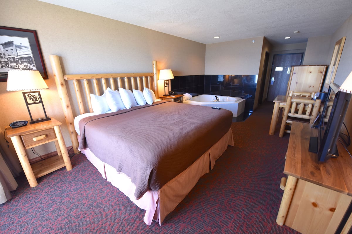 A hotel room with a large bed, a wooden headboard, a bedside table with a lamp, a small desk with a chair, a bathtub, and a mirror.