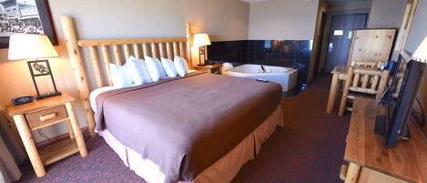 A hotel room with a large bed, a wooden headboard, a bedside table with a lamp, a small desk with a chair, a bathtub, and a mirror.