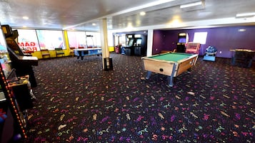 Ein Billardtisch in einem Raum mit bunten Bodenmustern, Spielautomaten und einem großen Fenster mit Blick auf die Umgebung.
