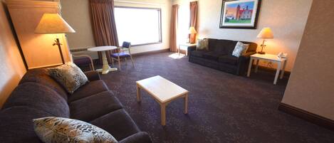 A hotel room with a dark brown sofa, a small white table, a dark brown armchair, two lamps, a framed picture on the wall, and a large window with curtains.