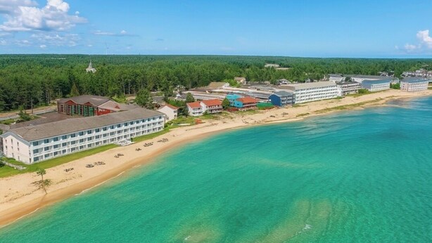 An aerial view of a coastal hotel with multiple buildings, a sandy beach, and clear turquoise waters. - Bridge Vista Beach - Hotel & Convention Center (Mackinaw City)
