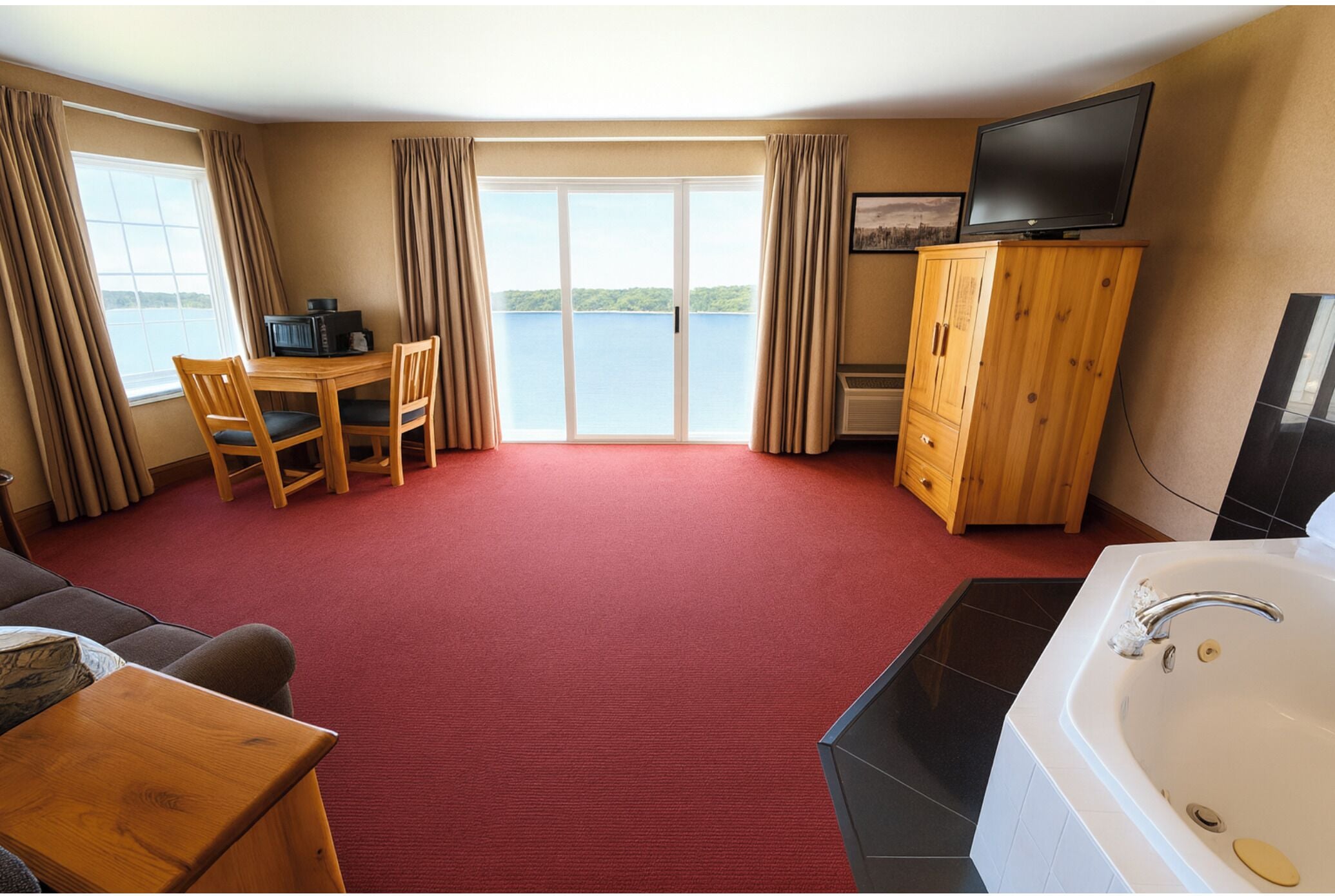 A hotel room with a red carpet, a wooden table with chairs, a TV on a wooden cabinet, a bathtub, and a large window with curtains.