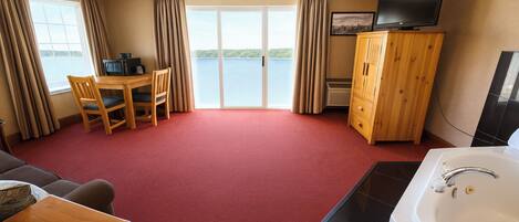 A hotel room with a red carpet, a wooden table with chairs, a TV on a wooden cabinet, a bathtub, and a large window with curtains.