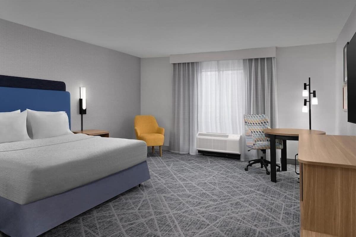 A hotel room with a bed, a desk, a chair, a window with curtains, and a radiator.
