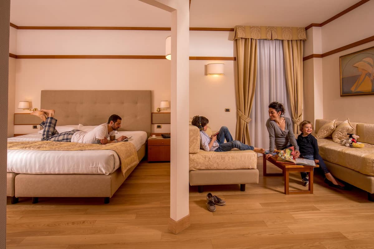 A hotel room with a large bed, a sofa, a coffee table, and a small table. There are lamps on the nightstands and a painting on the wall.
