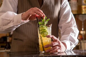 A person in a white shirt and vest preparing a drink with a straw and mint garnish.