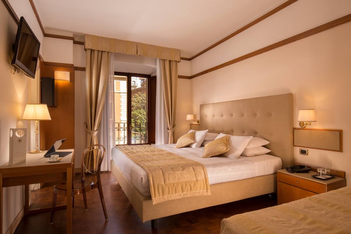 A hotel room with a large bed, bedside tables, a desk with a computer, and a chair.