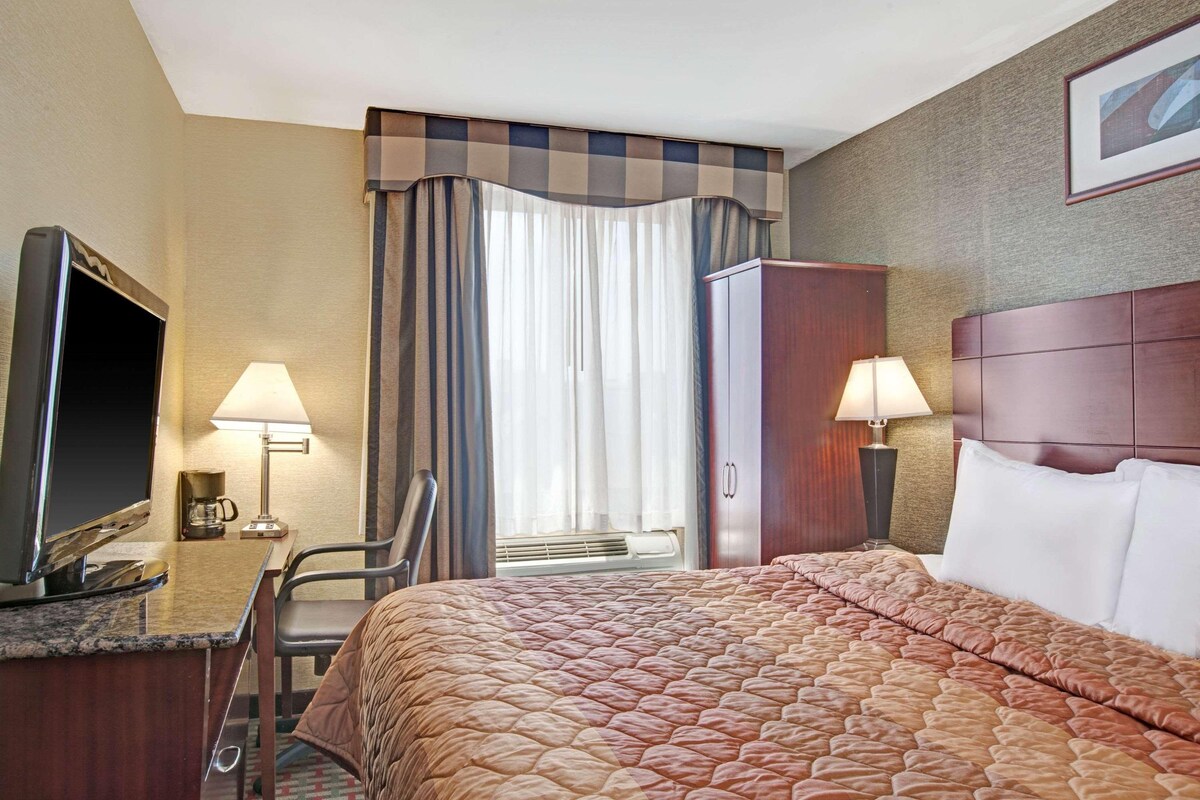 A hotel room with a bed, a desk with a television, two lamps, a chair, a wardrobe, and a window with curtains.