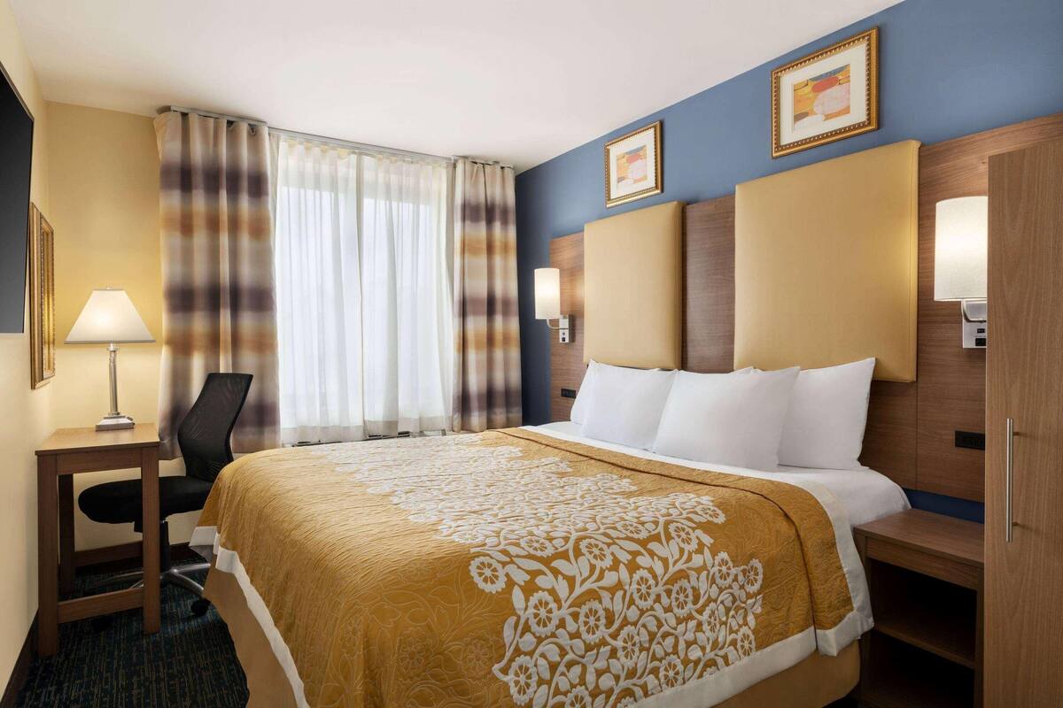 Habitación de hotel con cama, escritorio, silla, lámpara y ventana con cortinas.