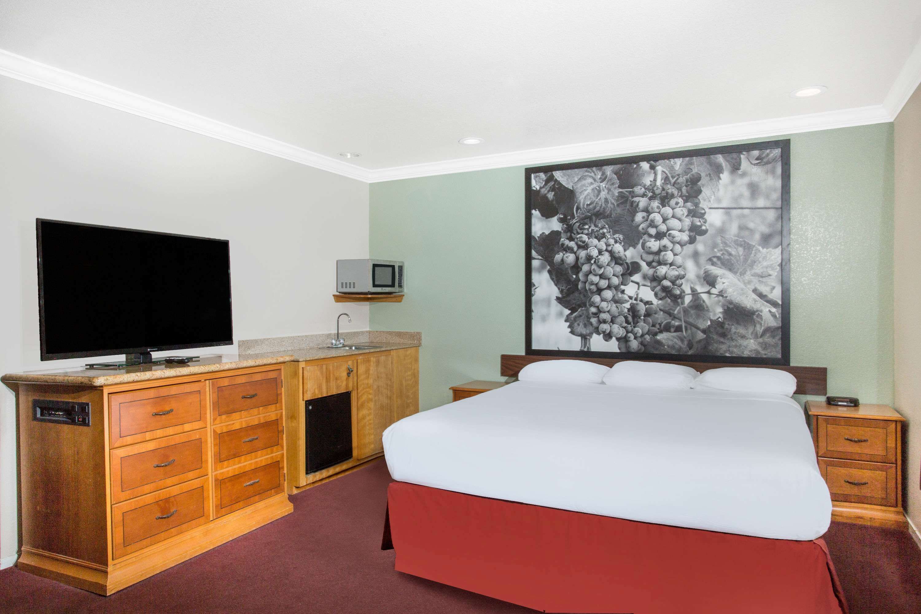 A hotel room with a bed, a TV, a wooden dresser, and a wall-mounted picture.