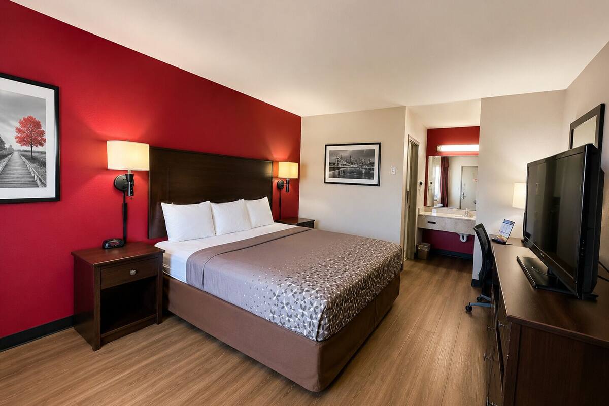 A hotel room with a bed, bedside lamps, a TV, a desk with a computer, and a bathroom with a mirror and sink.