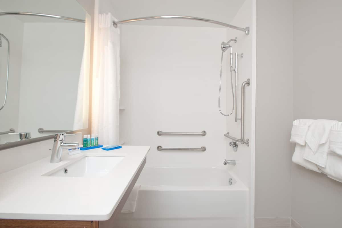 A modern bathroom with a white sink, mirror, and shower area. It includes a grab bar for safety and a towel rack.