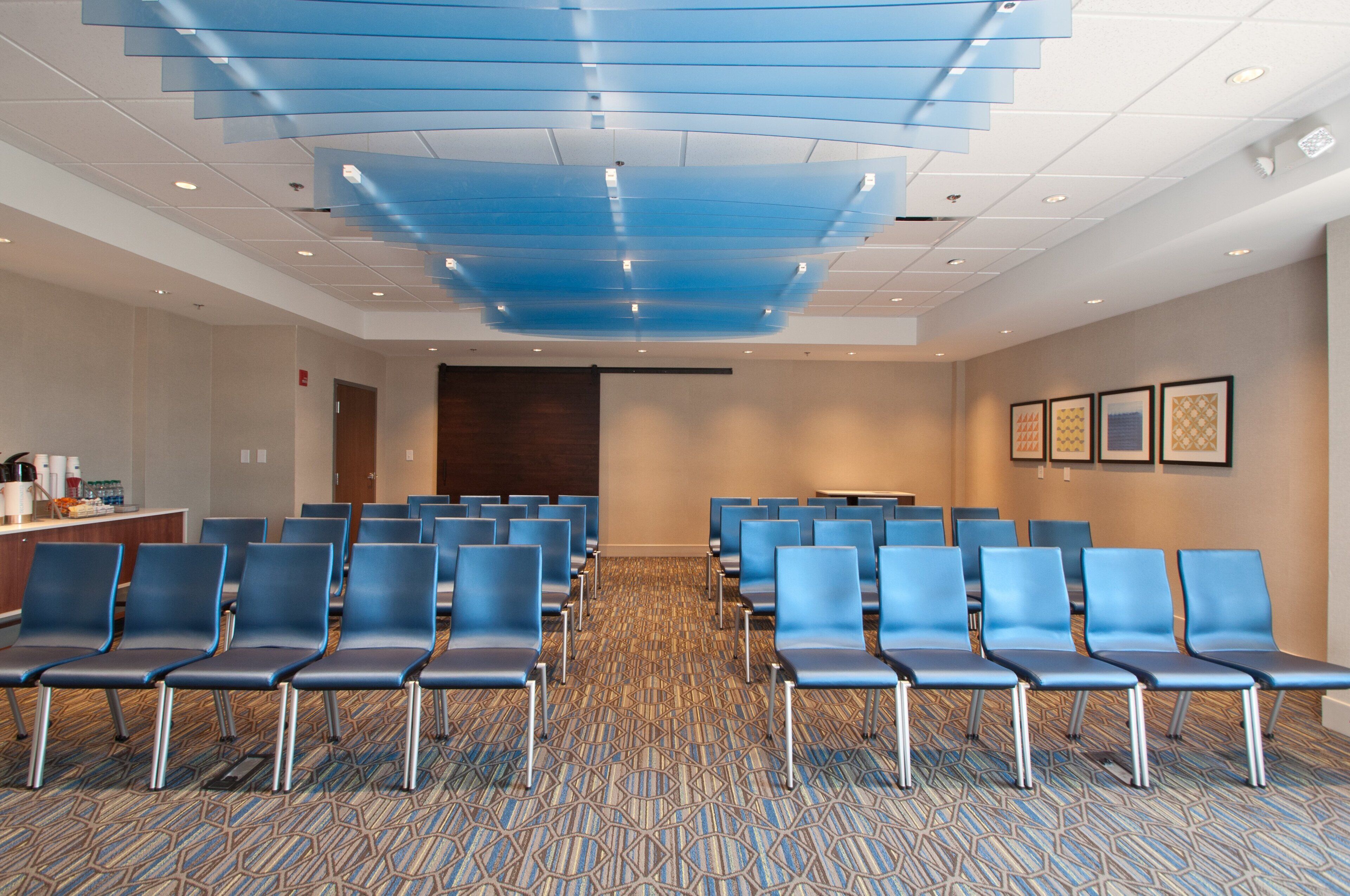 Un salón de conferencias con filas de sillas, piso alfombrado, techo con iluminación empotrada y cuadros en la pared.