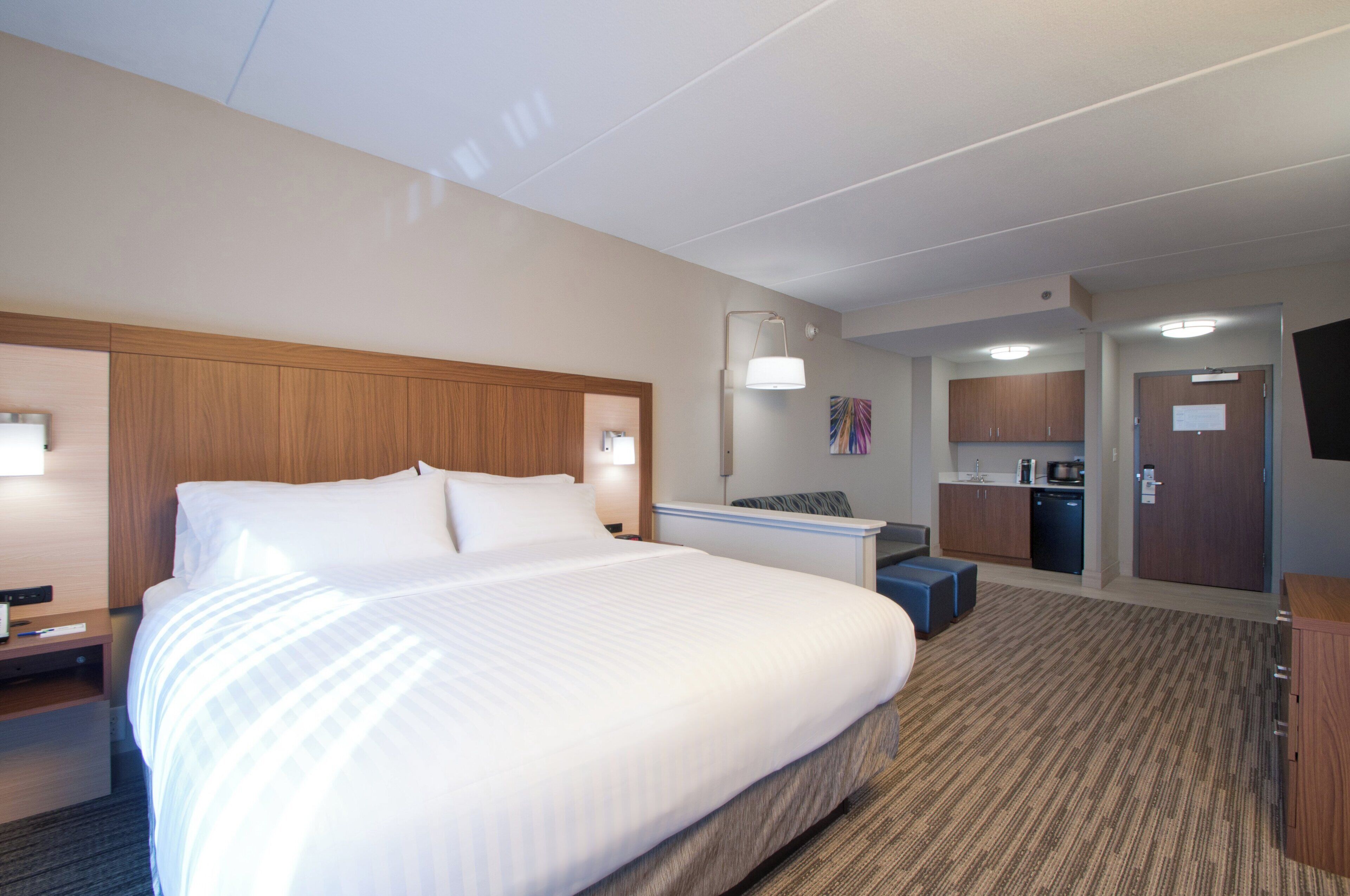 A hotel room with a large bed, a desk, a chair, a kitchenette, and a television.