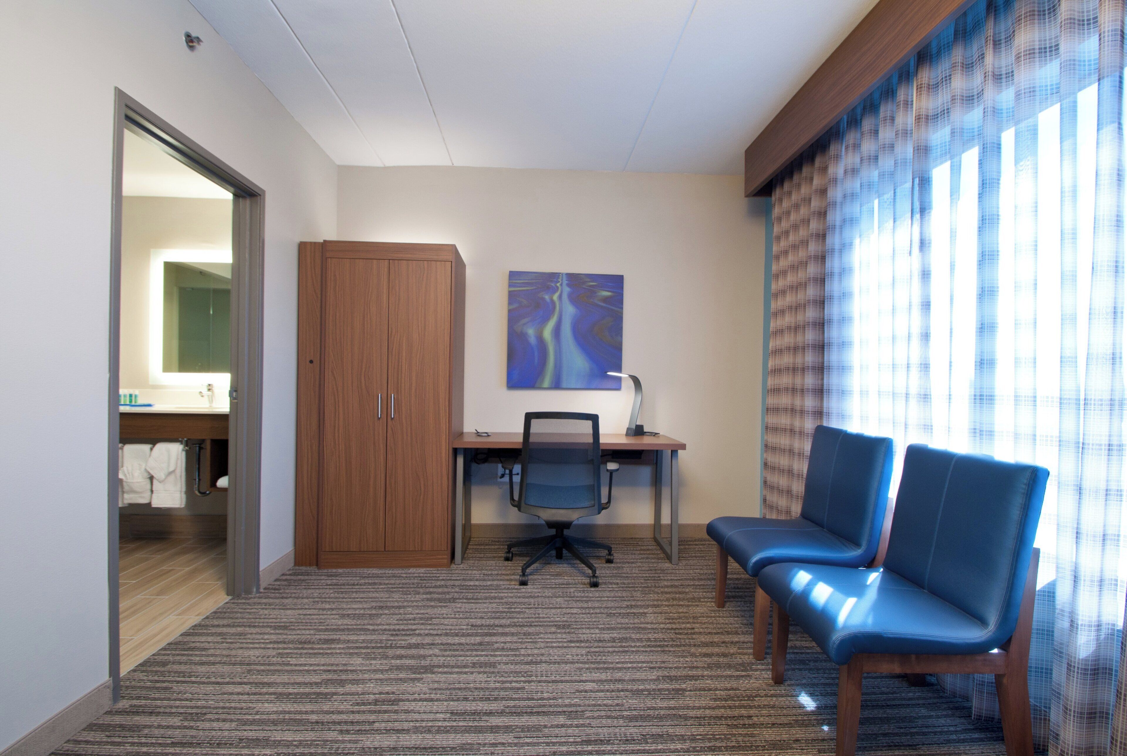 A hotel room with a desk, two chairs, a painting on the wall, and a bathroom visible through an open door.
