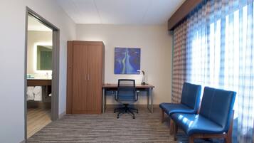 A hotel room with a desk, two chairs, a painting on the wall, and a bathroom visible through an open door.