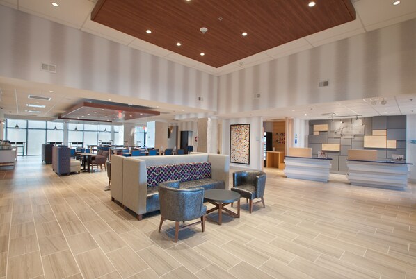 Un hall d’entrée spacieux au design moderne, comprenant un grand espace salon avec des canapés et des fauteuils, un comptoir d’accueil et, au fond, un coin cuisine.