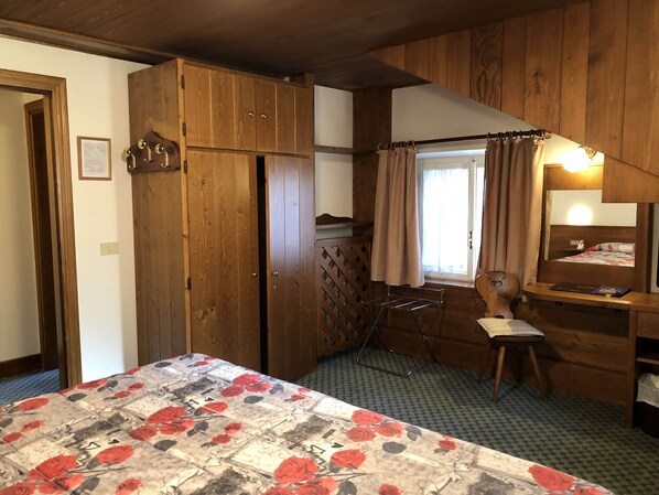 A bedroom with a wooden wardrobe, a bed with a floral bedspread, a small desk with a mirror, and a window with curtains. - Hotel Majoni (Cortina d'Ampezzo)