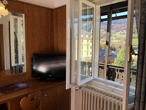 Una habitación con paredes de madera, un televisor sobre una mesa de madera, un ventanal con vistas a edificios y montañas, y un radiador debajo de la ventana.