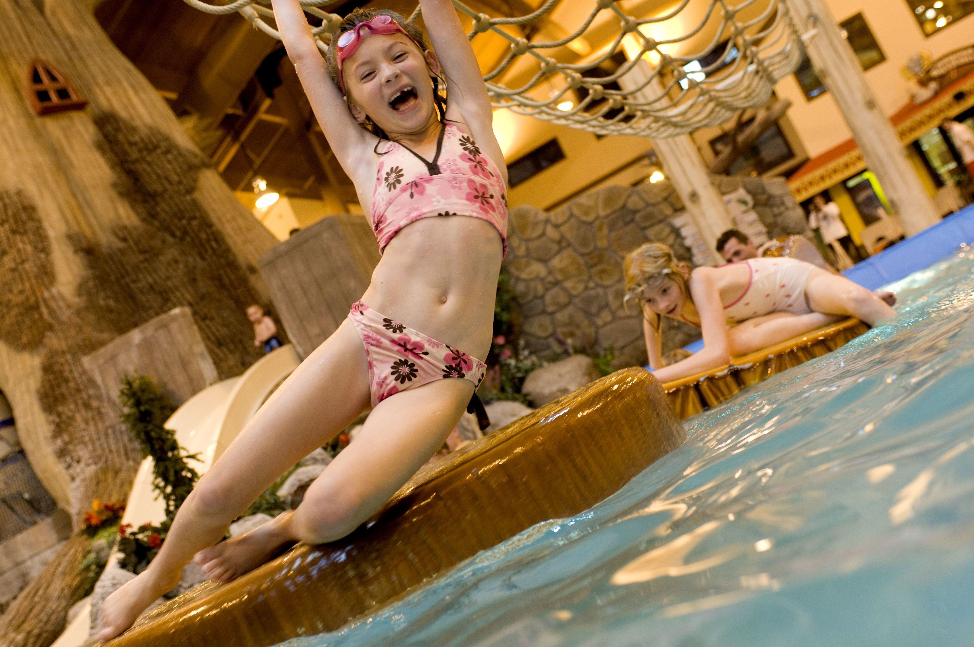 Una persona con traje de baño floral sobre una plataforma de madera en una zona de piscina, con paredes de piedra y un puente colgante.