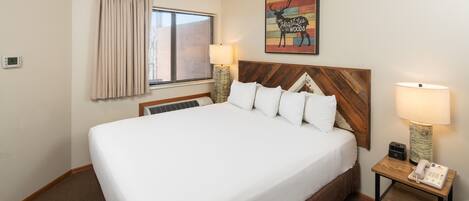 Habitación de hotel con una cama grande, cabecera de madera, mesita de noche con lámpara, una obra de arte en la pared y una ventana con cortinas.