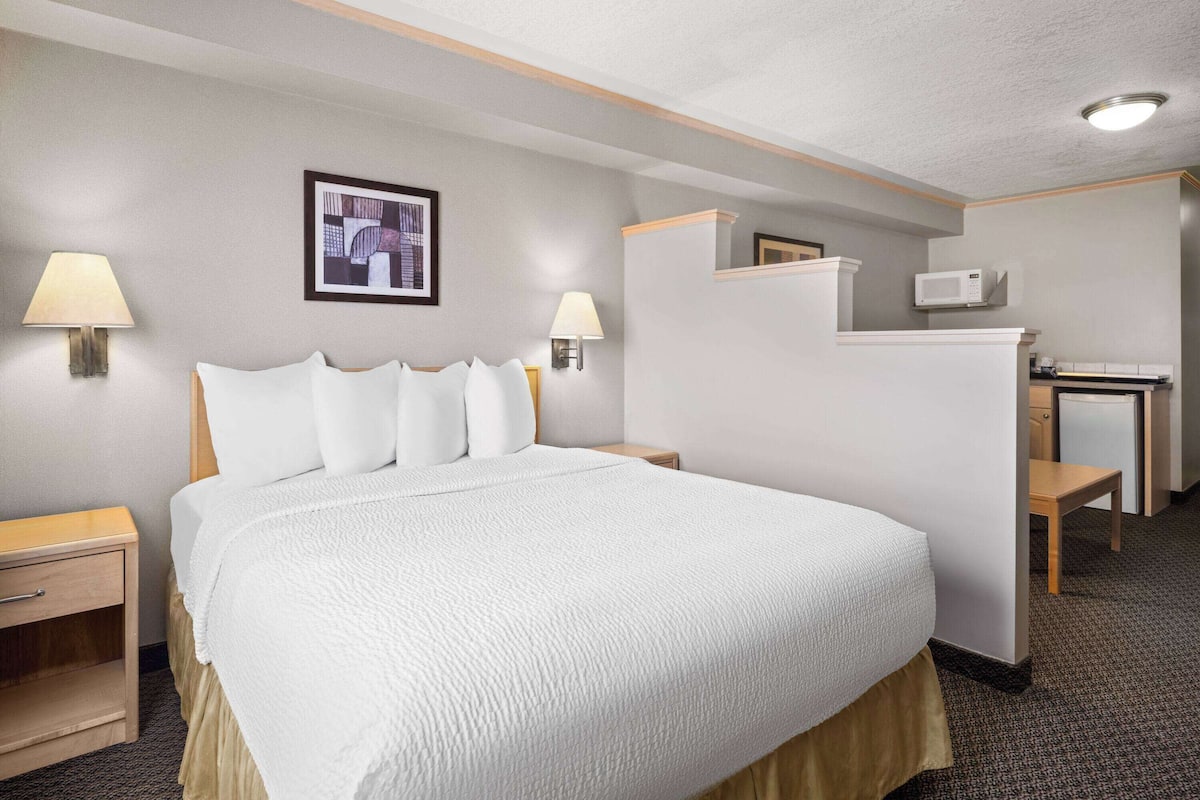 A hotel room with a large bed, two bedside lamps, a small desk with a chair, a microwave, and a framed picture on the wall.