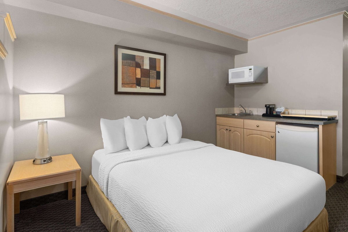 A hotel room with a bed, bedside table, lamp, artwork, microwave, and a small kitchenette.