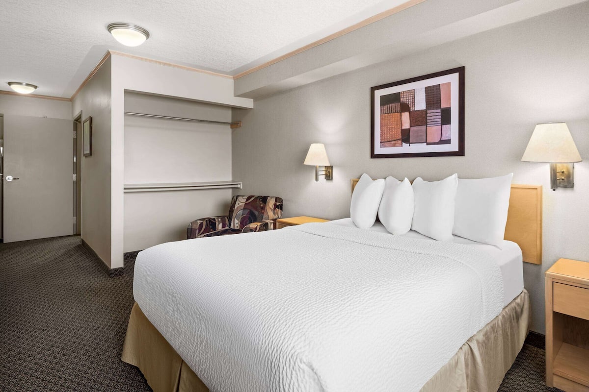 A hotel room with a large bed, a chair, a desk, and a wall-mounted lamp.