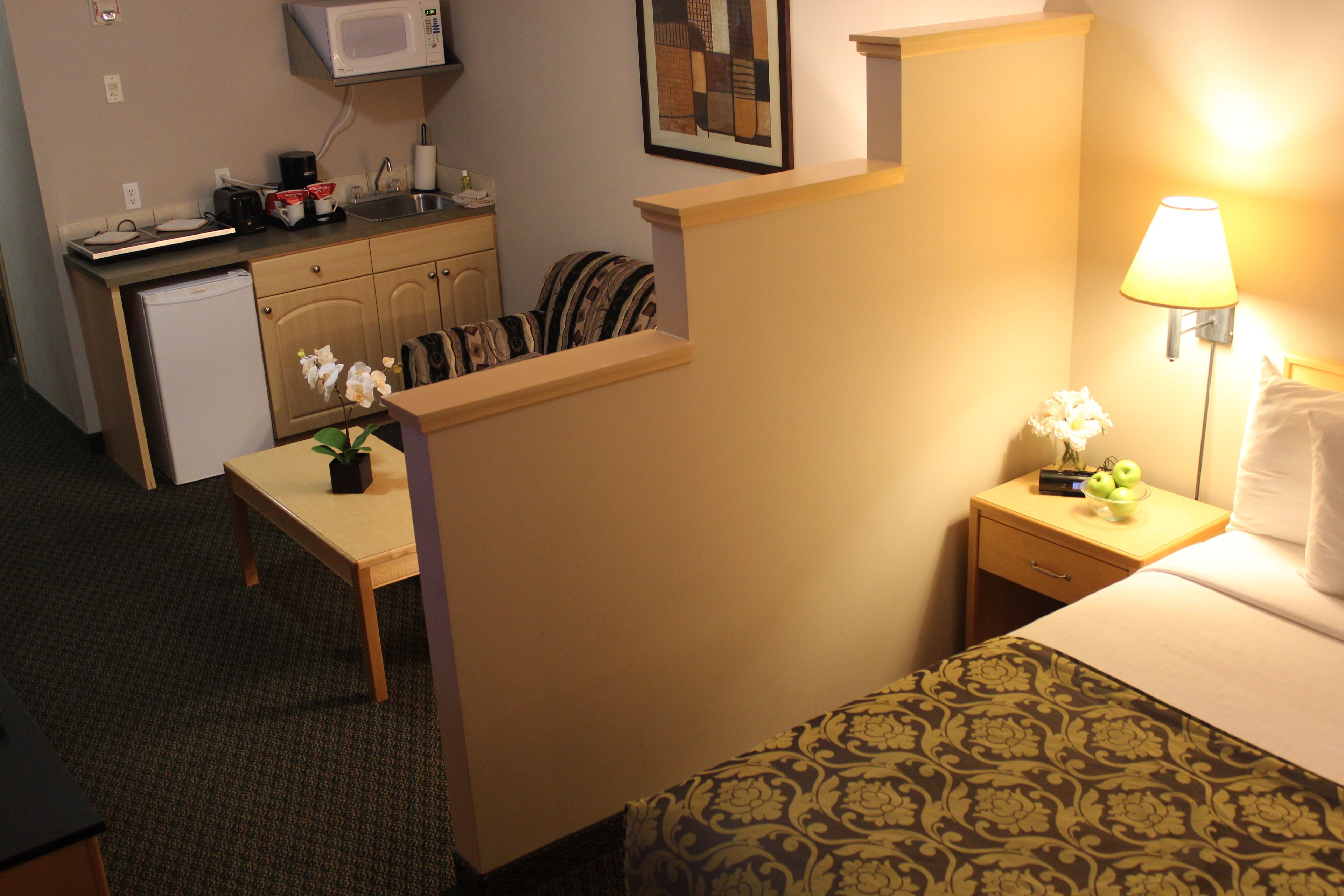 A hotel room with a bed, a bedside table with a lamp, a small desk with a computer, a microwave, and a refrigerator.