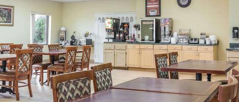 A dining area with wooden tables and chairs, a kitchen area with coffee machines, and a clock on the wall.