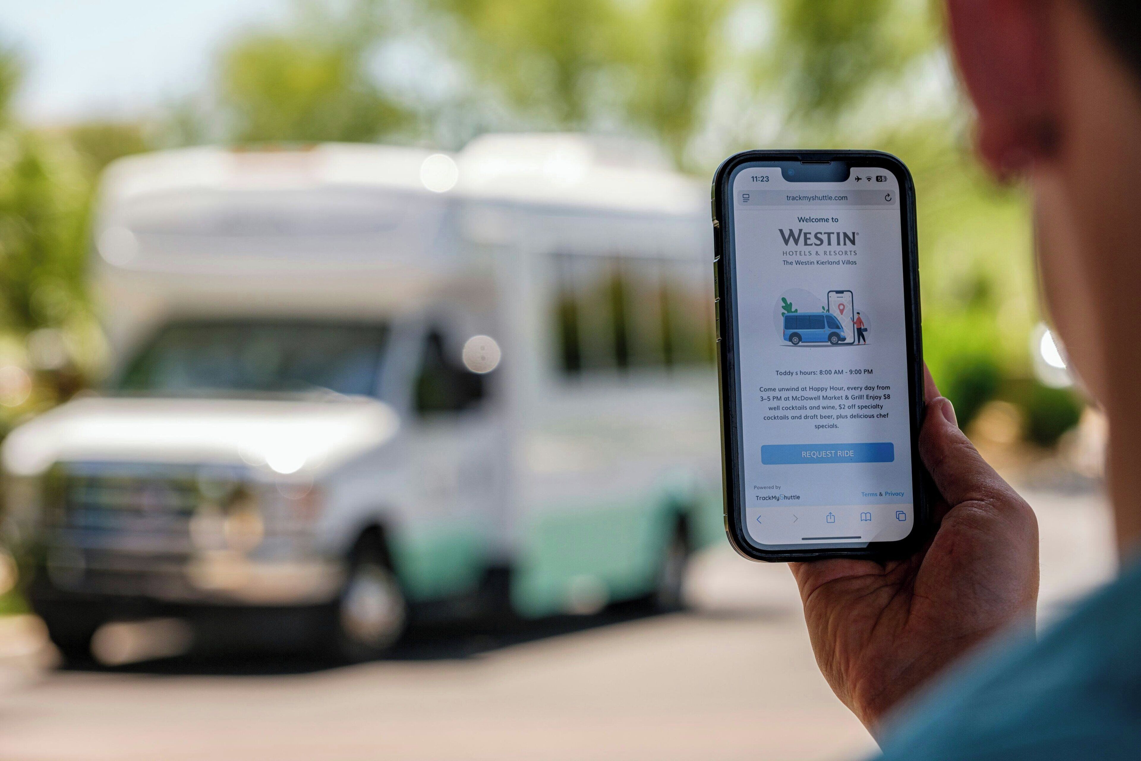 Eine Person hält ein Smartphone, auf dem eine Hotelbuchungs-App angezeigt wird, im Hintergrund ist ein Fahrzeug zu sehen.