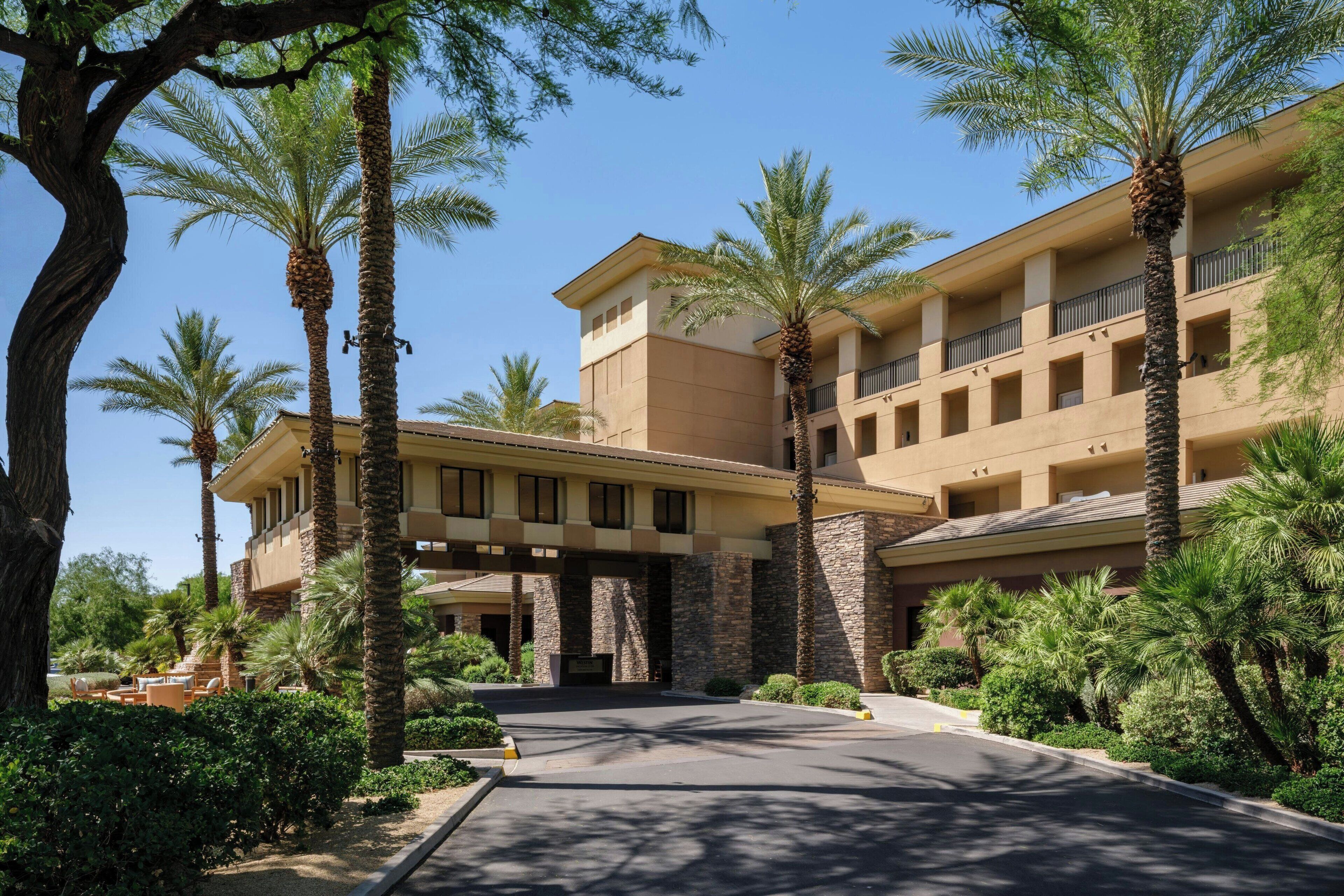 A multi-story hotel with a beige facade, palm trees, and a paved driveway.