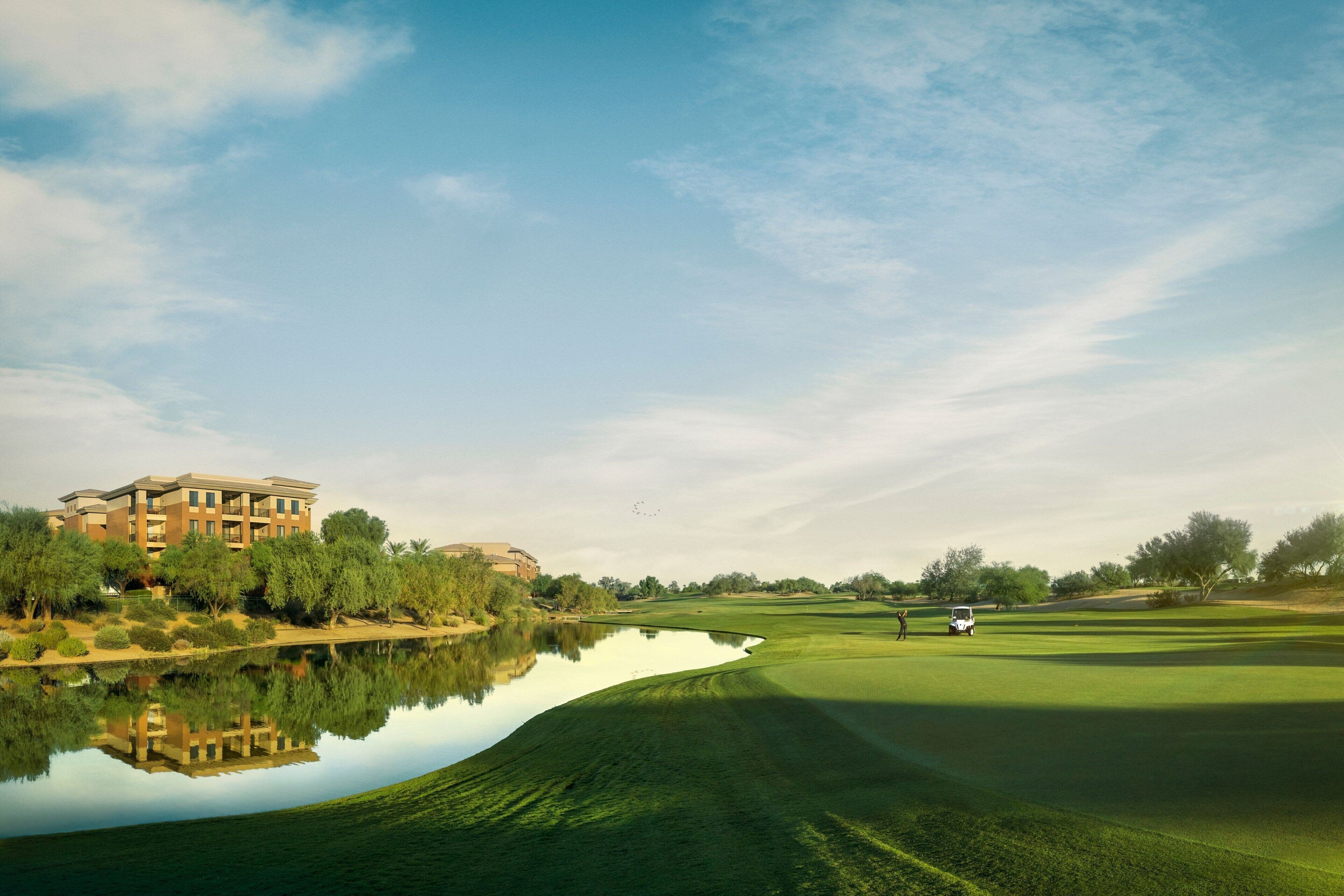 A golf course with a pond reflecting a building, trees, and a clear sky.