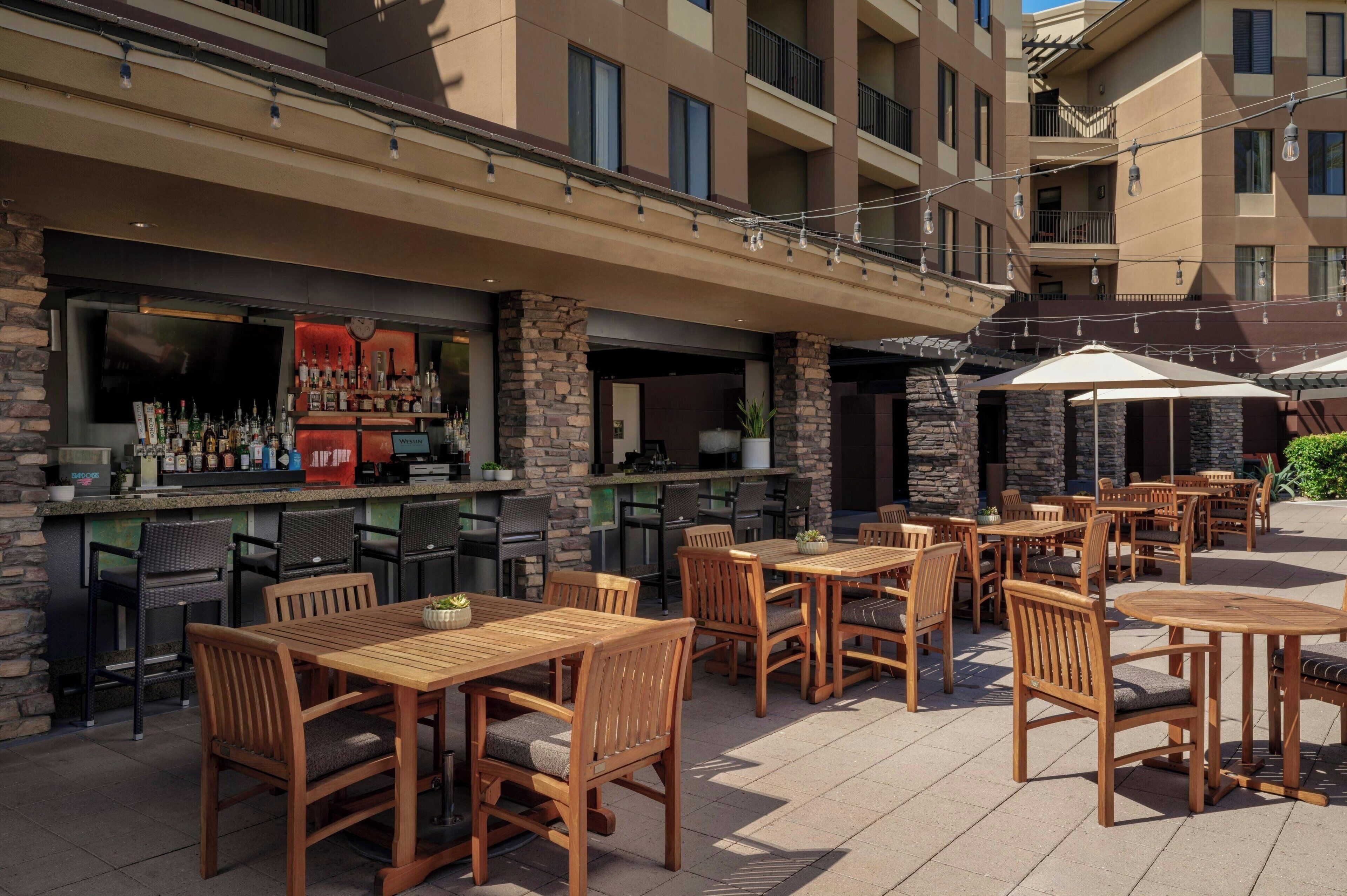 An outdoor patio with wooden tables and chairs, a bar area with bottles, and a stone facade.