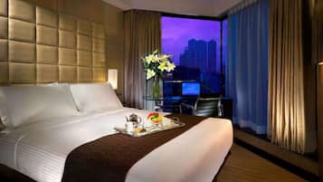 A hotel room with a large bed, a desk, and a view of a cityscape at dusk.