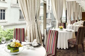A dining area with striped chairs and tables, a vase of flowers, and a view of a building through large windows. - The Kowloon Hotel (Kowloon)