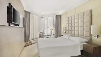 A hotel room with a large bed, a flat-screen TV mounted on the wall, a desk with a chair, and a bedside table with a lamp.