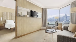 A hotel room with a bed, a seating area with a chair, a TV mounted on the wall, and a large window offering a city view. - The Kowloon Hotel (Kowloon)