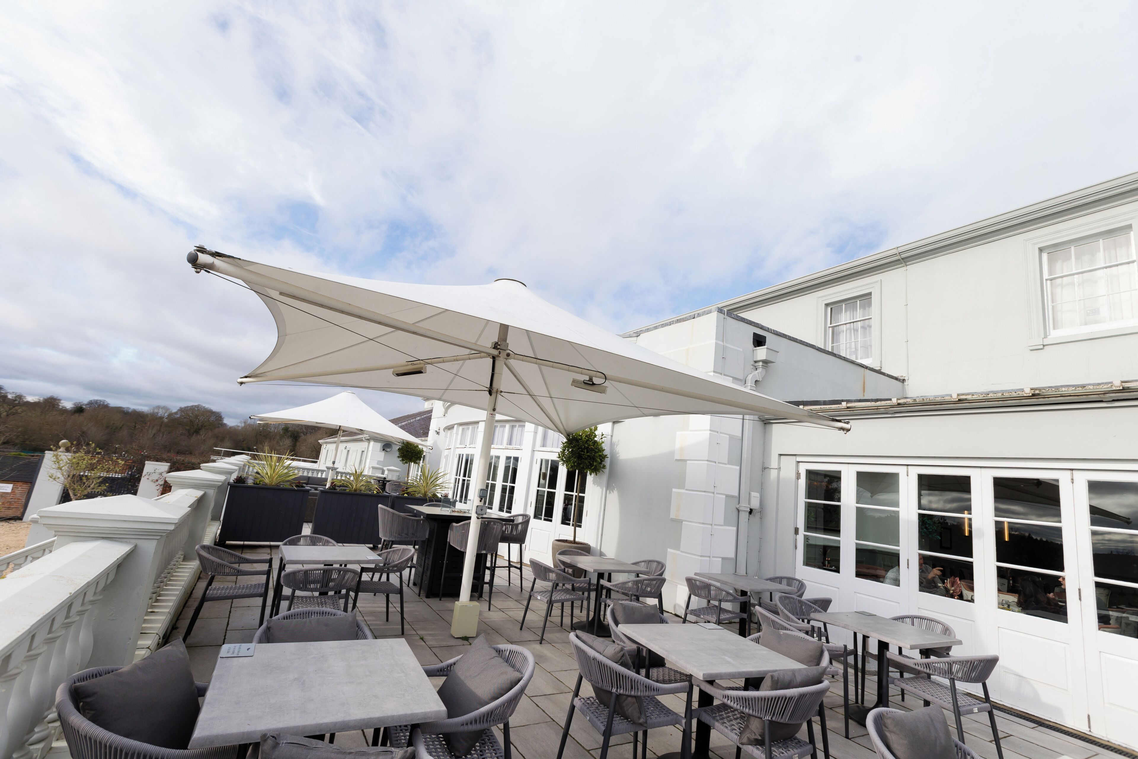 An outdoor patio with tables and chairs, a white umbrella, and a building with large windows.