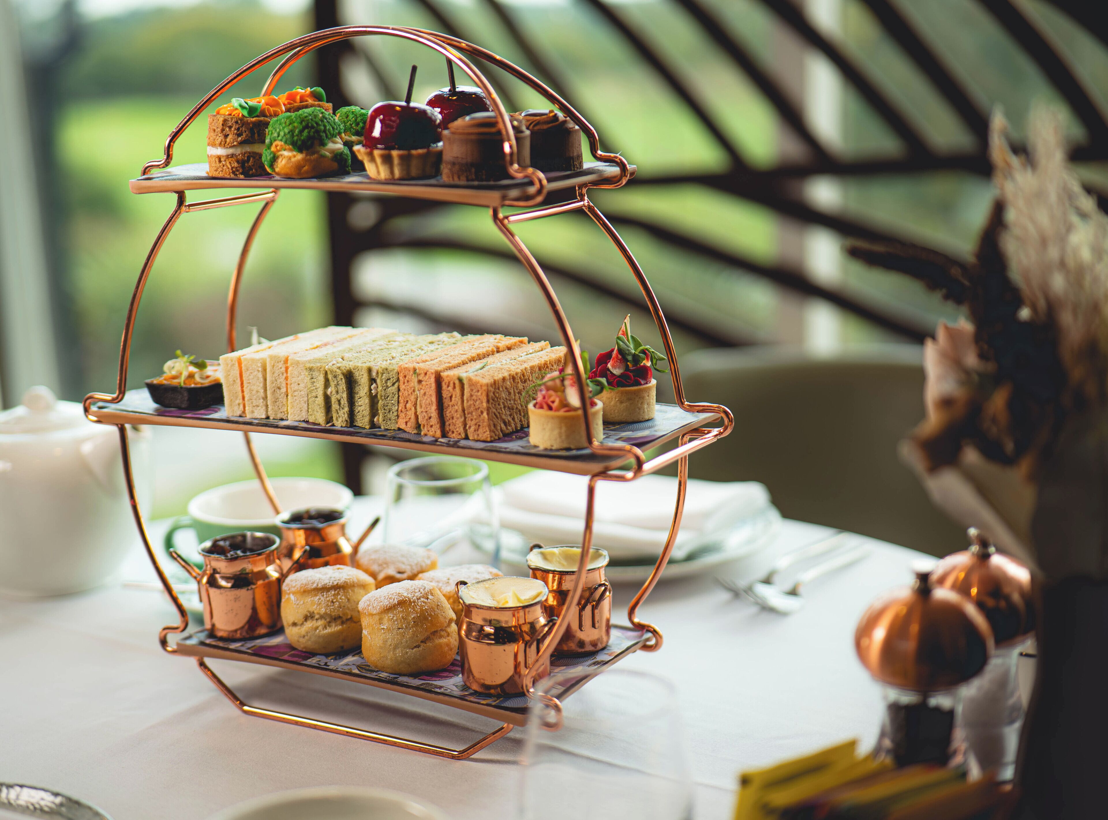 Un expositor escalonado para postres con sándwiches, pasteles y una tetera sobre una mesa.