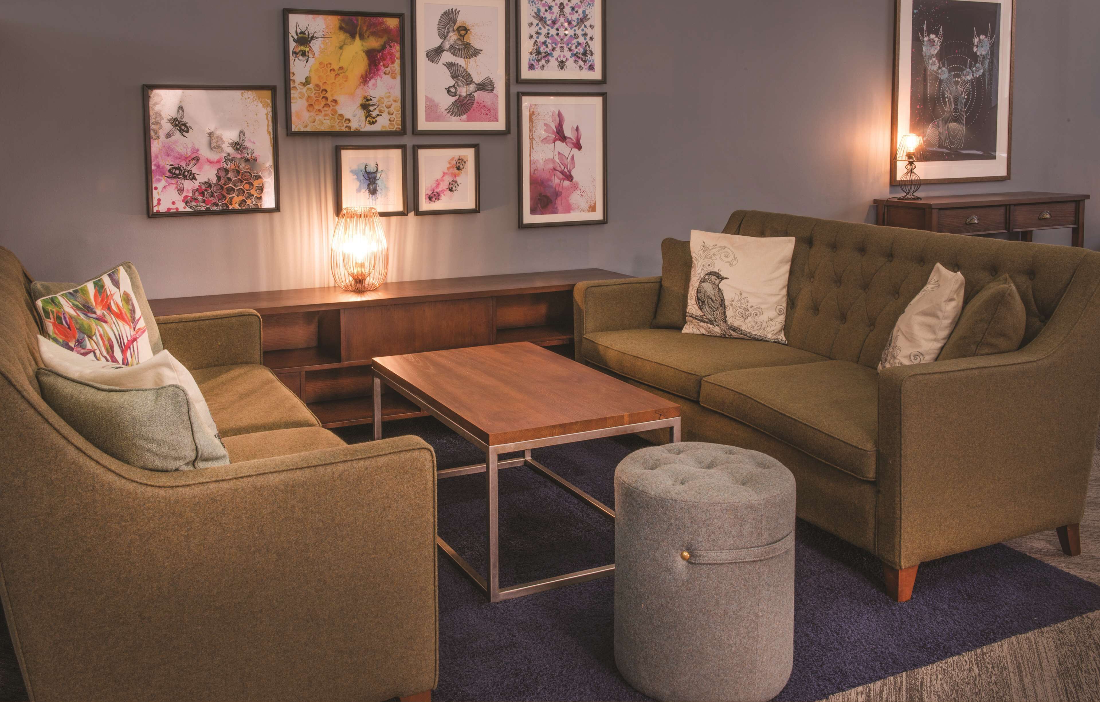 Un salon comprenant un canapé, un fauteuil, un pouf et une table basse. Des œuvres d’art encadrées ornent le mur, et une lampe se trouve sur la table d’appoint.