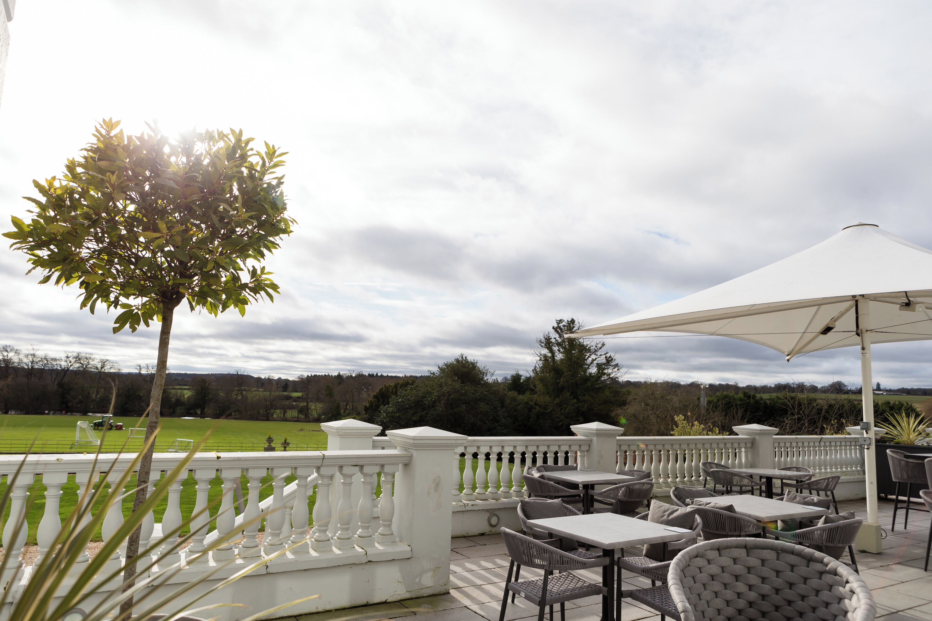 An outdoor patio area with tables and chairs, a white umbrella, and a view of a green field.