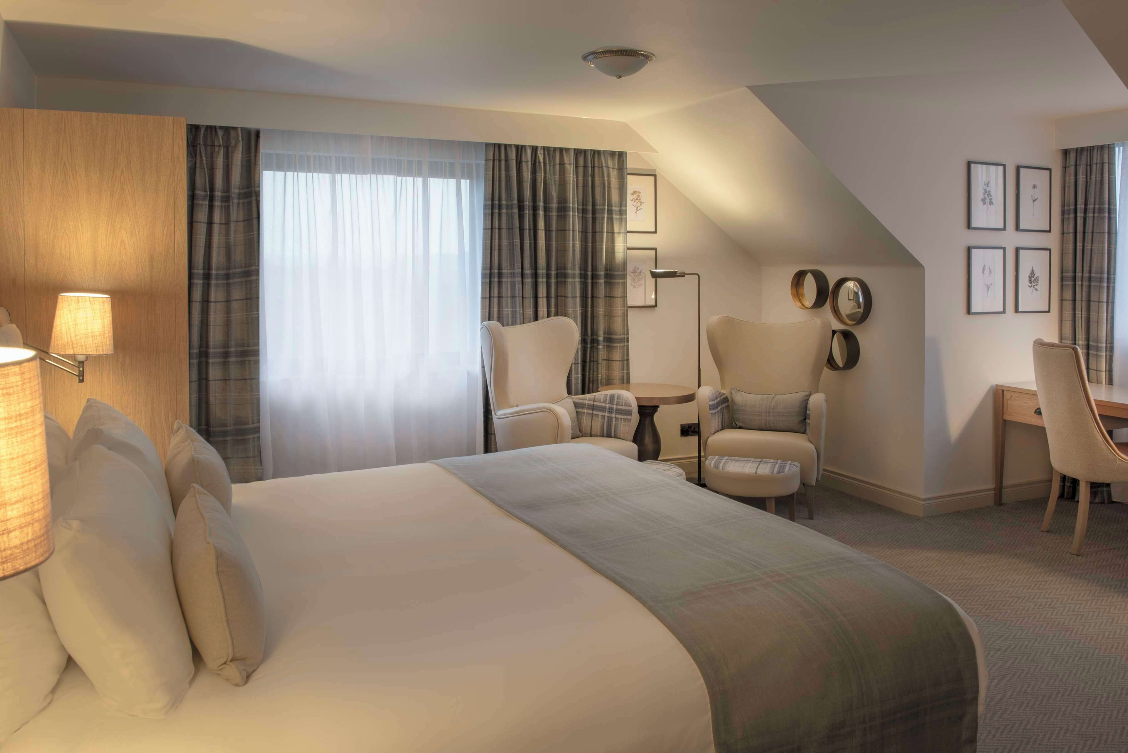 Habitación de hotel con una cama grande, un escritorio, una silla y una mesita. Hay cortinas, una lámpara y cuadros en la pared.