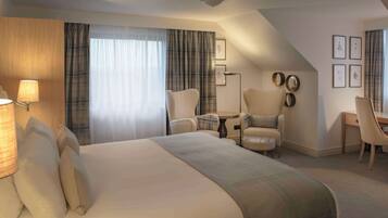Habitación de hotel con una cama grande, un escritorio, una silla y una mesita. Hay cortinas, una lámpara y cuadros en la pared.