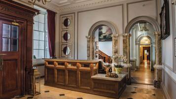 A grand lobby with a marble floor, wooden reception desk, and classical architecture including columns and arches.