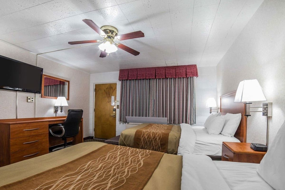 Una habitación de hotel con cama, escritorio y silla, televisor, ventilador de techo y ventana con cortinas.