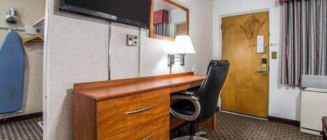 A hotel room with a wooden desk, a black office chair, a flat-screen TV mounted on the wall, a mirror above the desk, a lamp, a door with a wooden frame, and a patterned carpet.